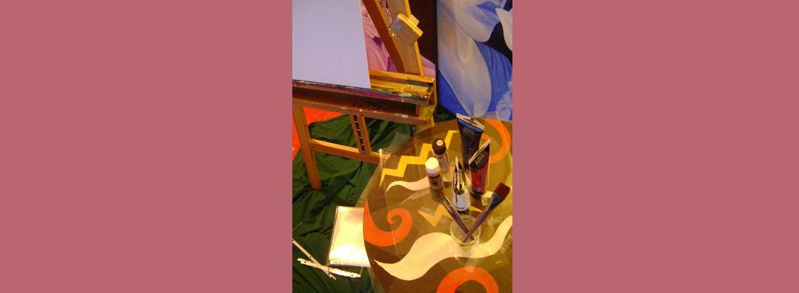 Photo shows an easel, a painted table with paints on it, and some paintings in the background